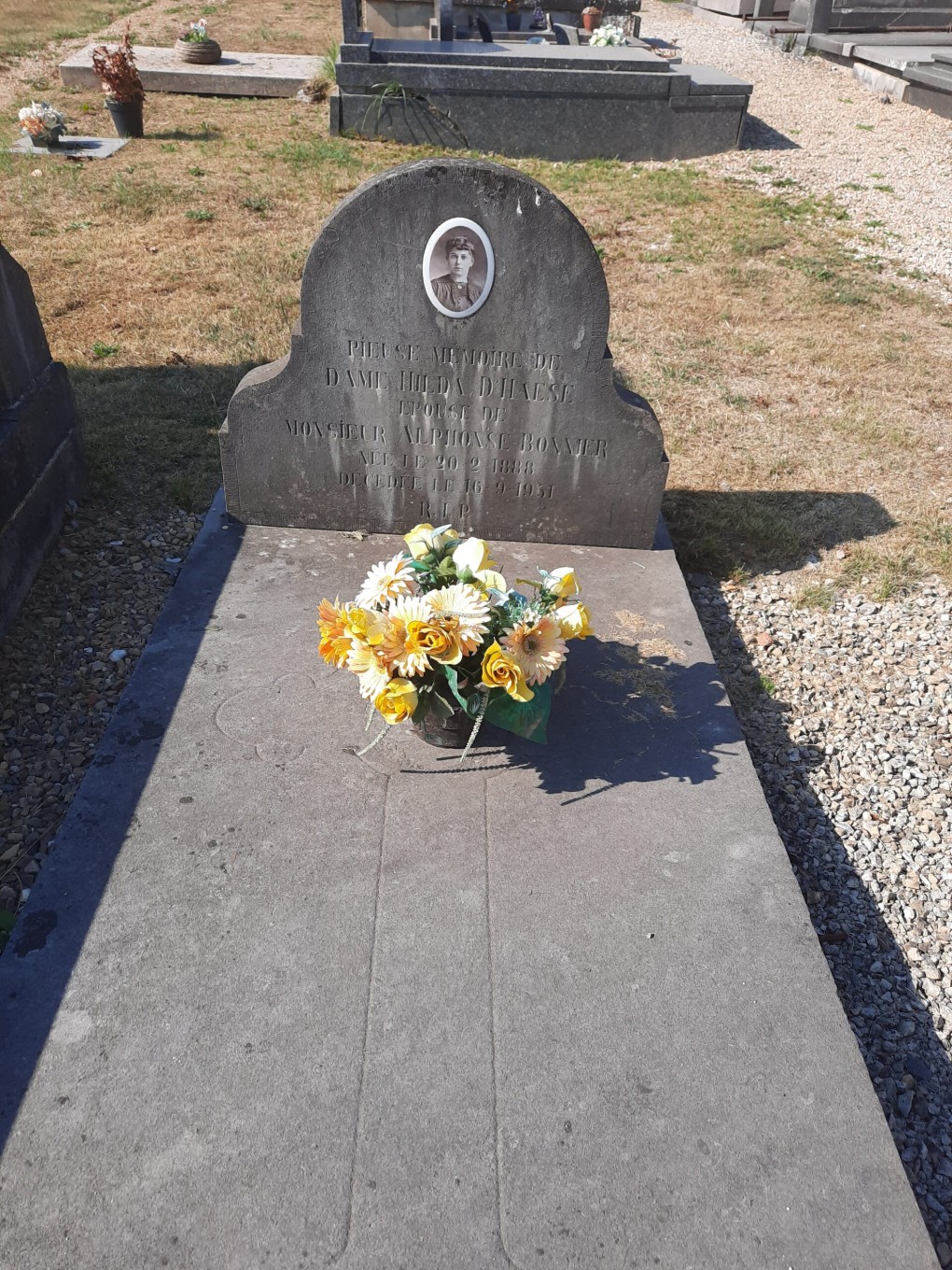 Grafsteen met foto en tekst, versierd met een boeket gele bloemen op een begraafplaats.