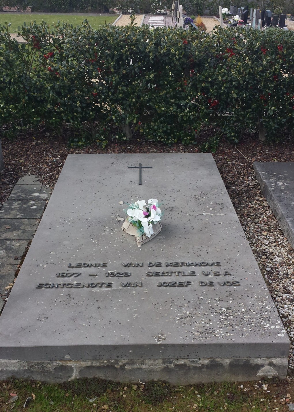 Grafsteen met tekst en bloemen, omringd door struiken met rode bessen op een begraafplaats.