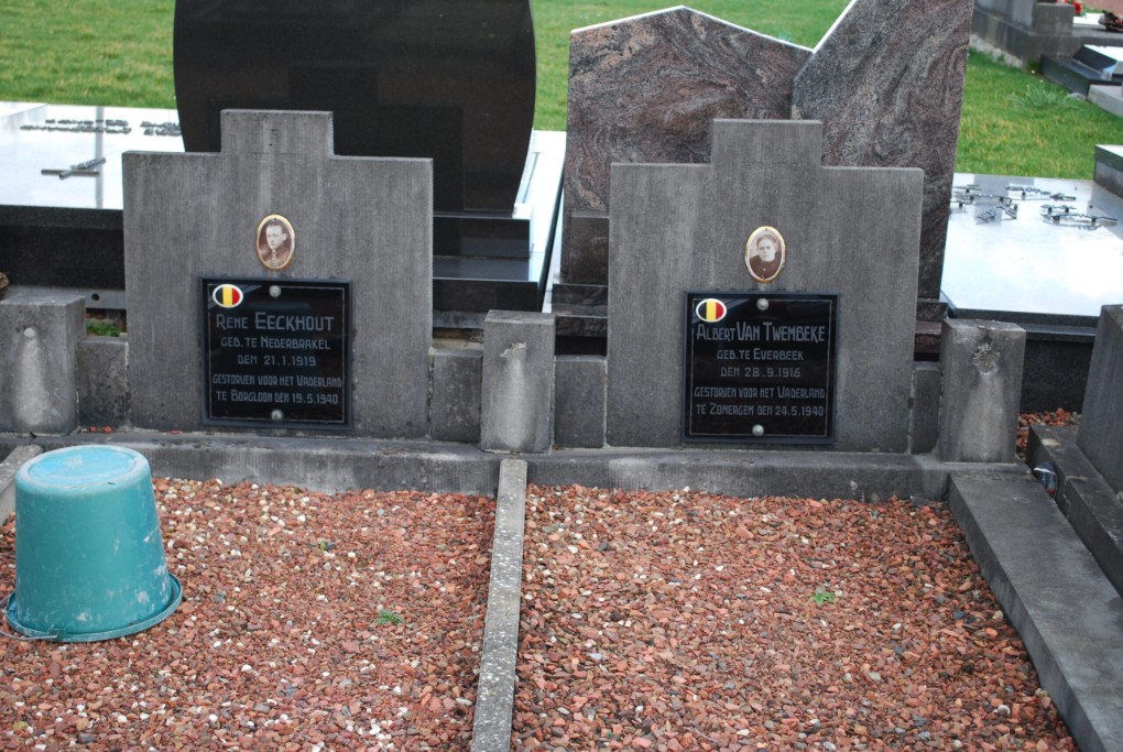 Twee graven met Belgische vlaggen en foto's van mannen, omgeven door rood grind en een groene emmer.