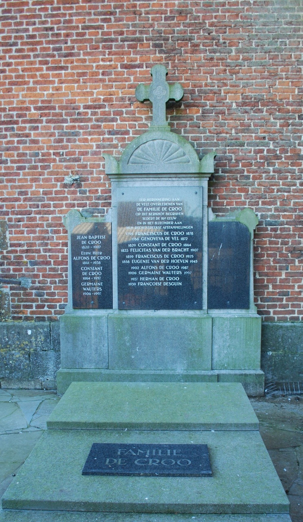 Grafsteen van de familie De Croo met namen en data, tegen een bakstenen muur, met een kruis bovenaan.