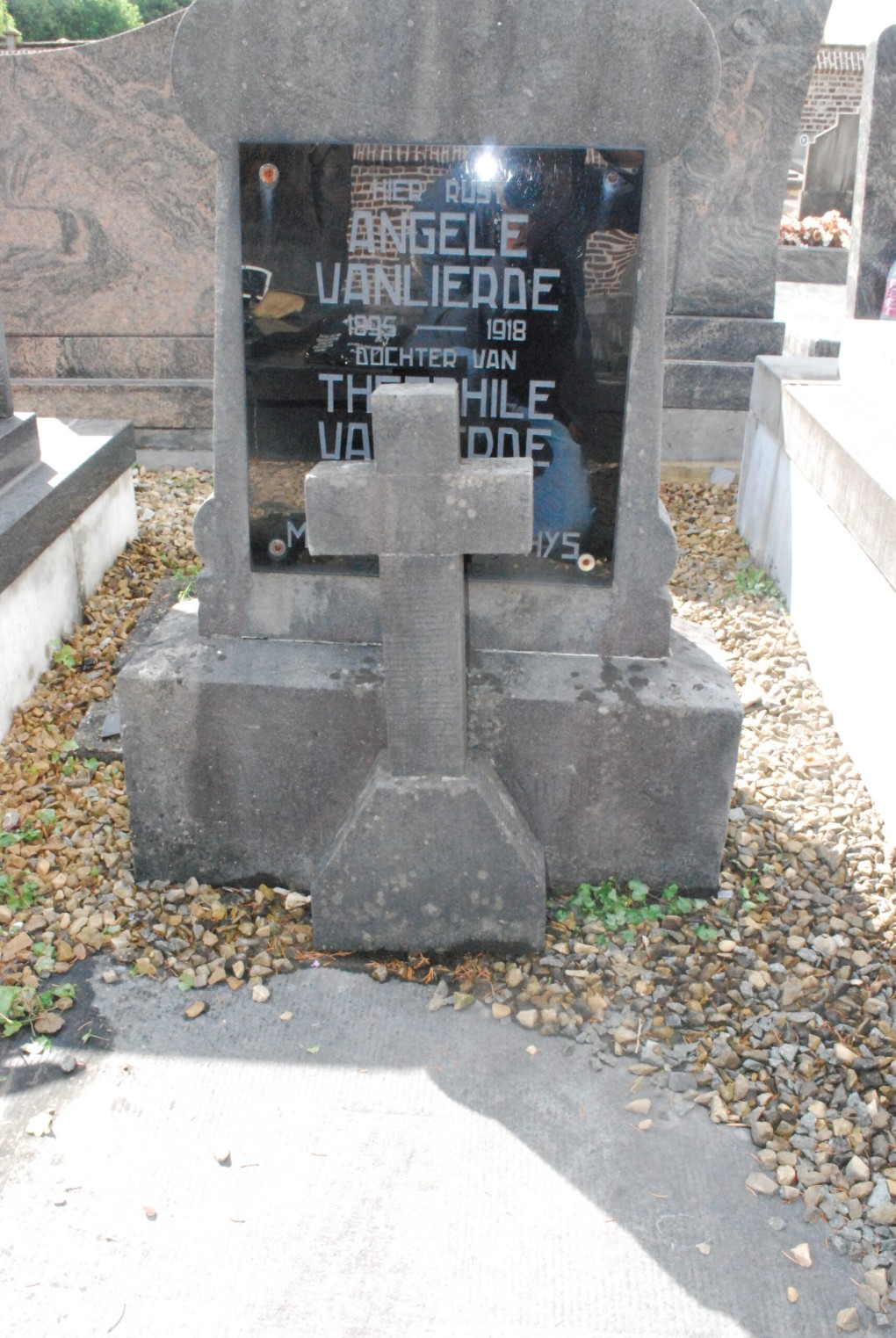 Grafsteen van Angele Vanlierde (1895-1918) met een stenen kruis ervoor, omgeven door grind en andere grafstenen.
