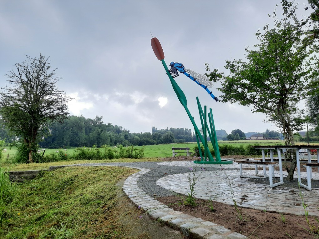 Beeld van een grote blauwe libelle op een kunstmatige rietstengel in een groene, landelijke omgeving.