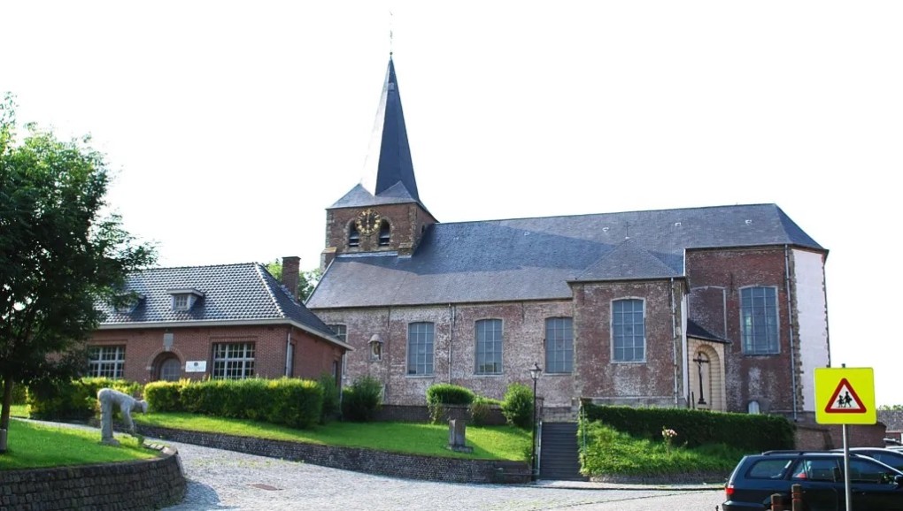 Kerkgebouw met hoge toren en klok, naast een huis met dakpannen, omgeven door groen en een verkeersbord.