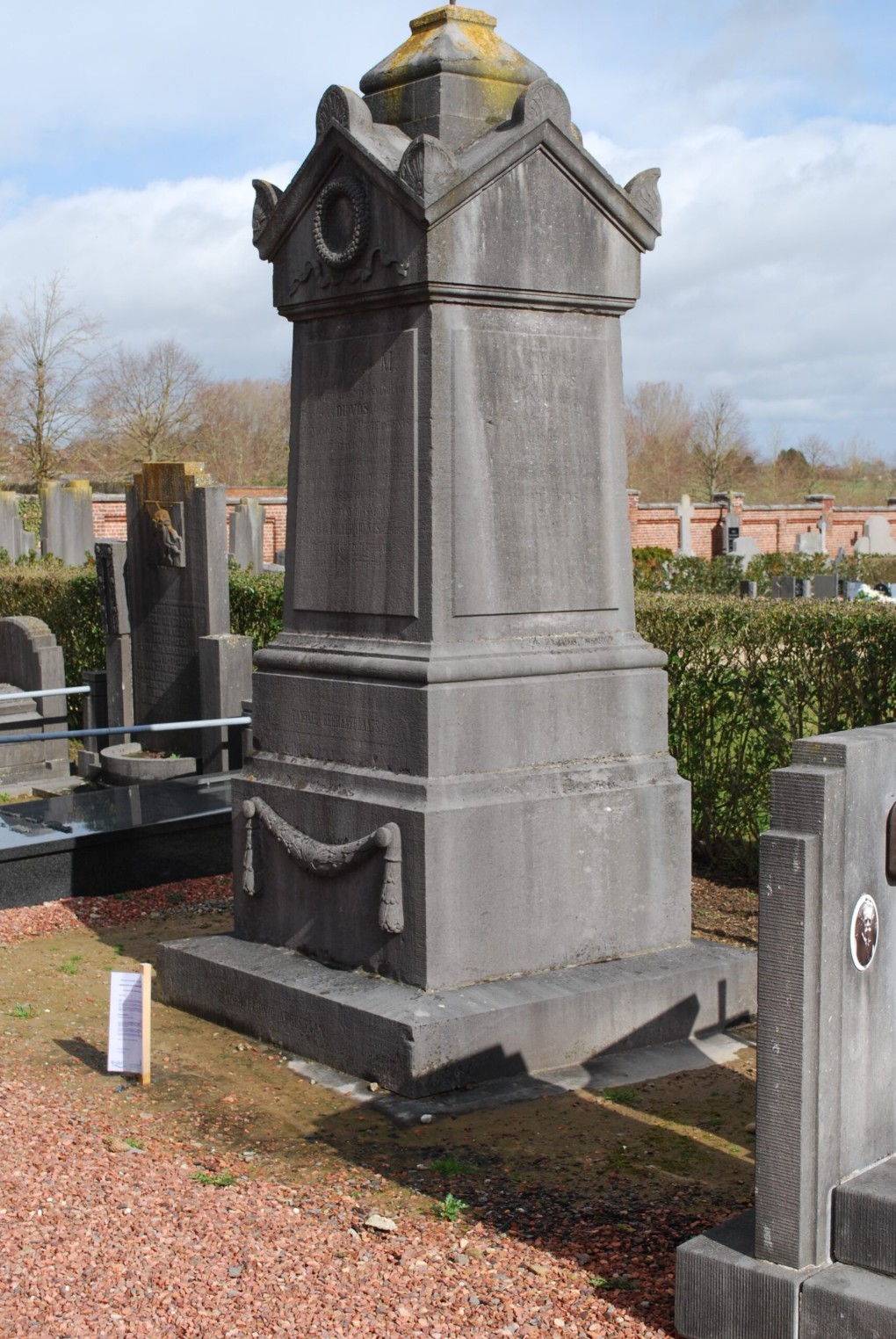 Groot grijs grafmonument met versieringen op een begraafplaats bij bewolkte hemel.