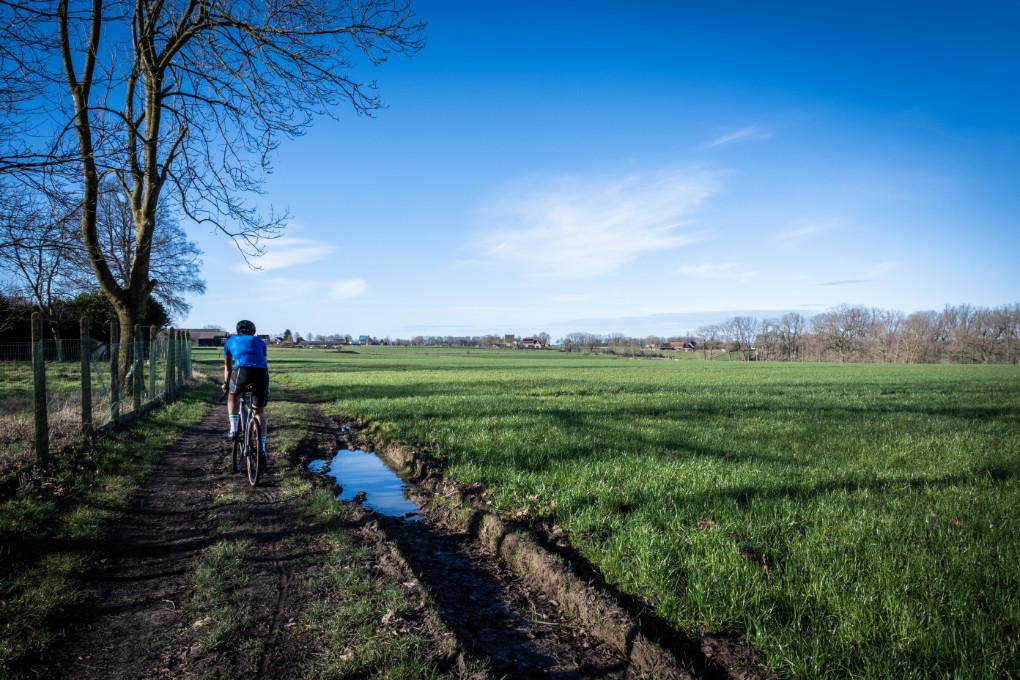 Fietser rijdt op een modderig pad langs een weiland onder een heldere blauwe lucht.
