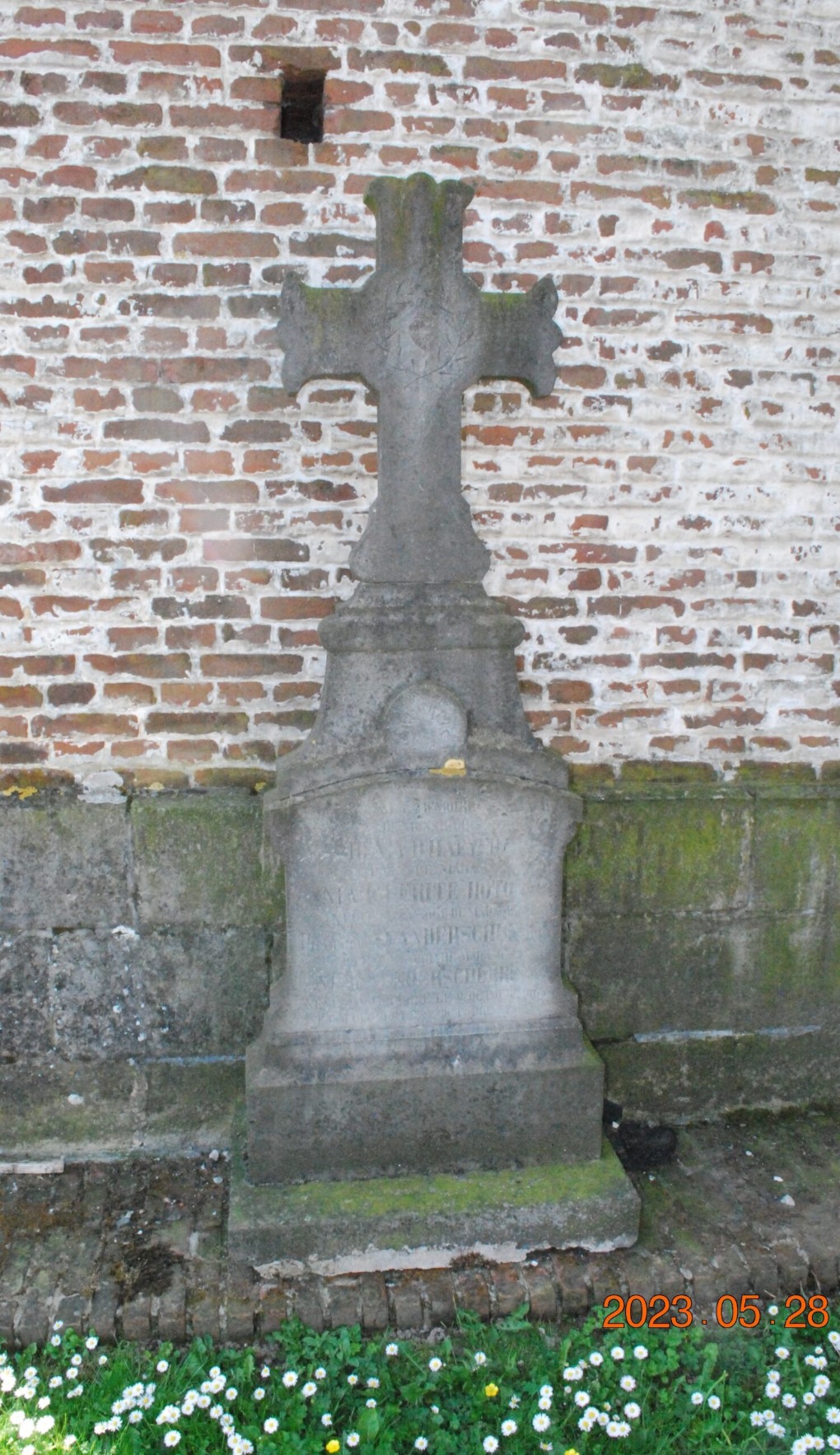 Oud grafmonument met kruis tegen een bakstenen muur, omgeven door gras en bloemen.