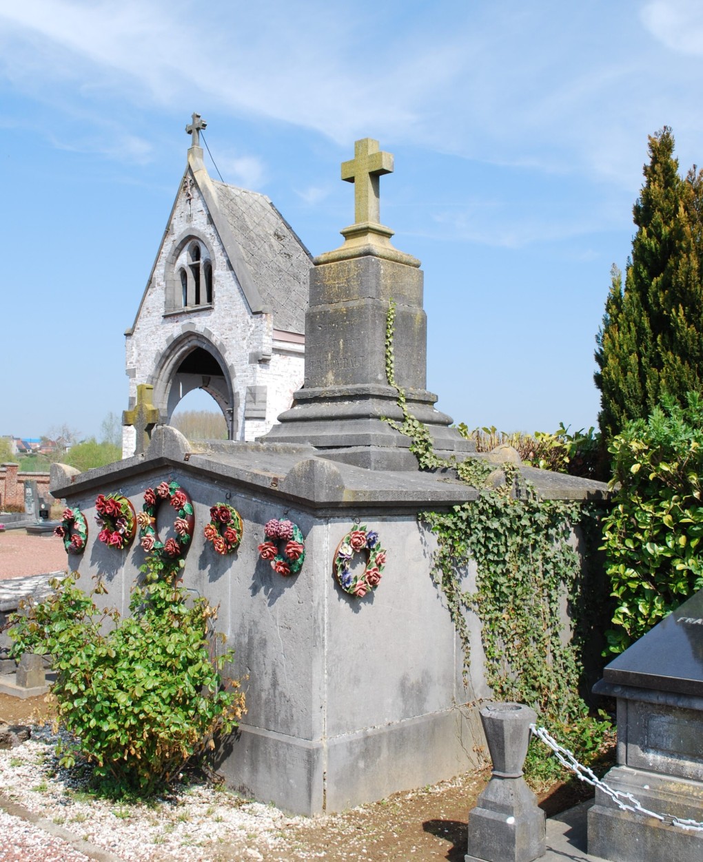 Grafmonument met kruis en bloemenkransen bij een kapel onder een blauwe lucht.