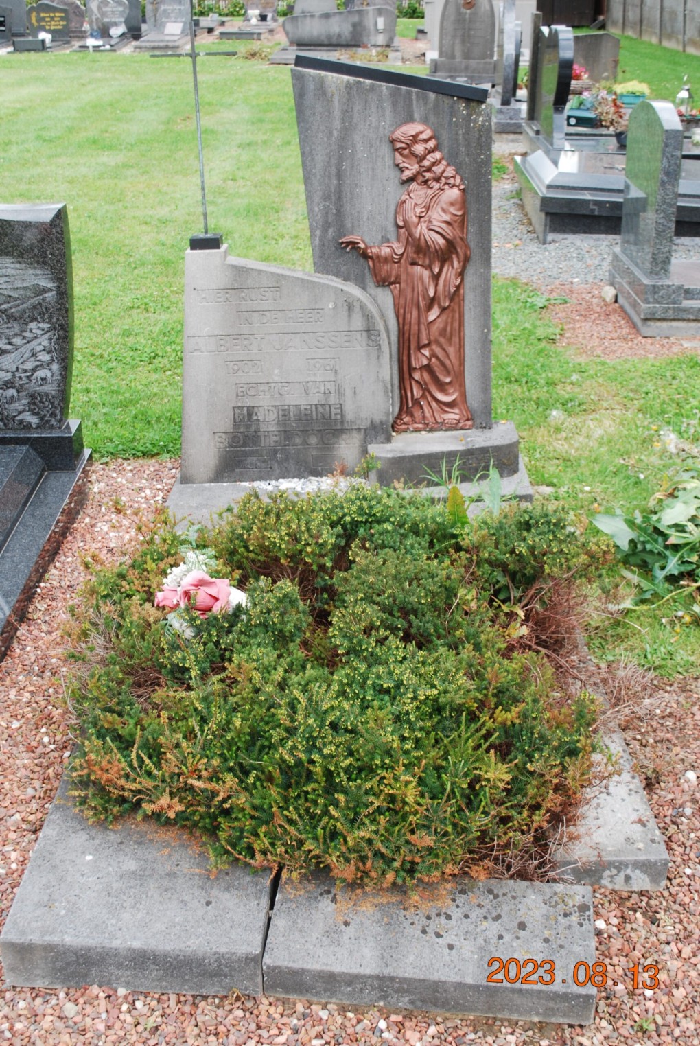 Grafsteen met een bronzen beeld van een biddende Jezus en begroeide begraafplaats.