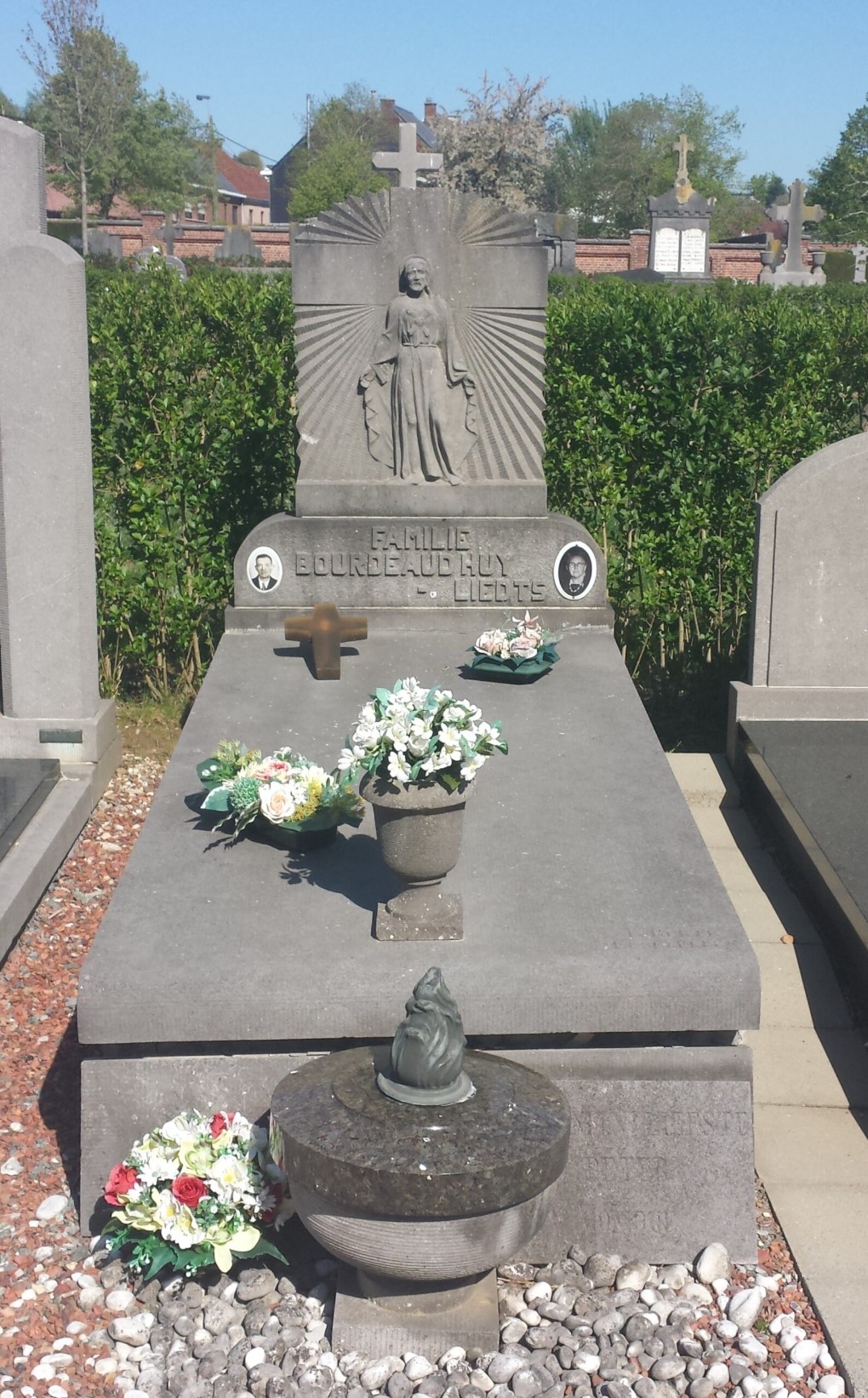 Grafsteen met beeld van Jezus en bloemen, familie Bourdeaudhuys-Liedts, omgeven door groen en andere graven.