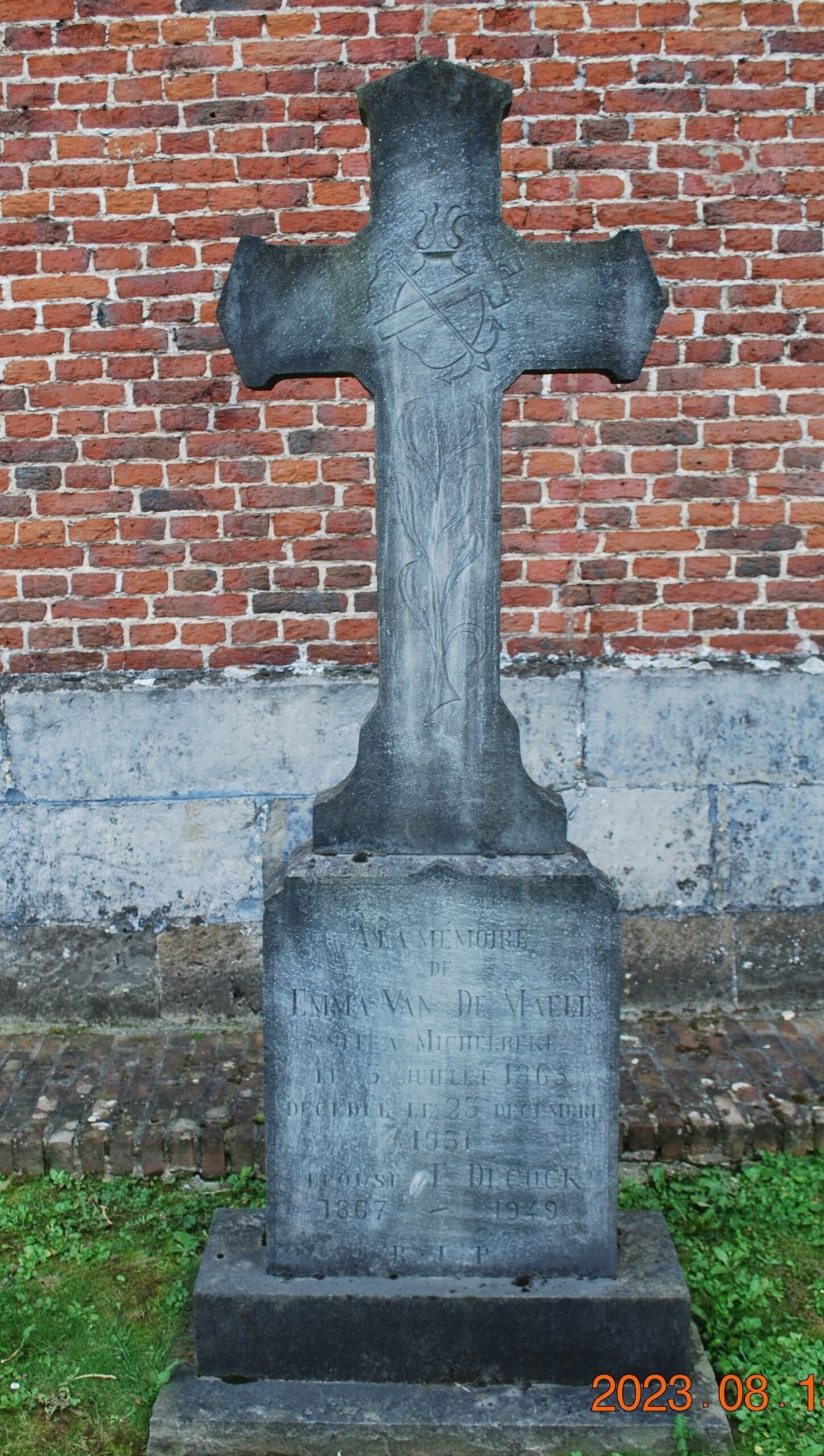 Grafsteen met kruis en inscriptie, geplaatst voor een bakstenen muur met gras ervoor.