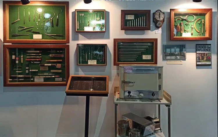 Museumwand met ingelijste medische instrumenten en een kleine oven op een tafel eronder.
