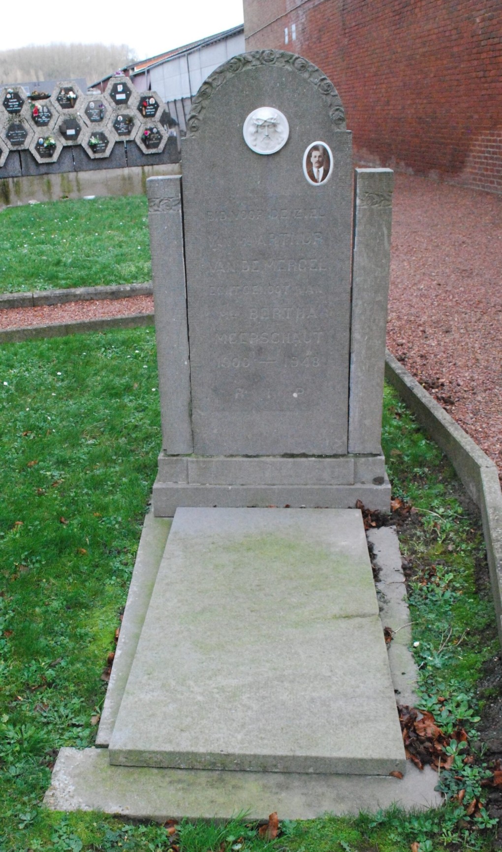 Grafsteen van Arthur Van De Mergel (1900-1948) met een foto en reliëf van een gezicht, omgeven door gras en andere grafstenen.