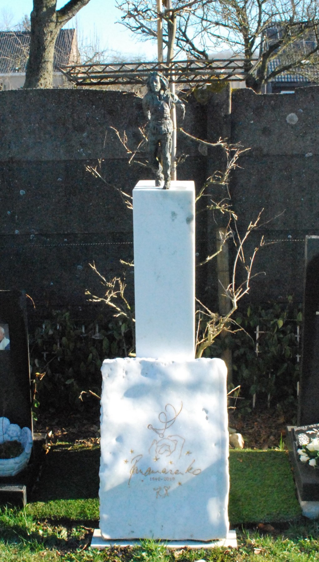Grafsteen met een witte zuil en een bronzen beeldje van een persoon bovenop, omgeven door bomen en andere graven.
