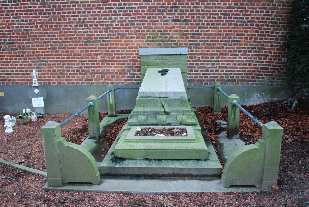 Oud grafmonument van groene steen met omheining, tegen een rode bakstenen muur.