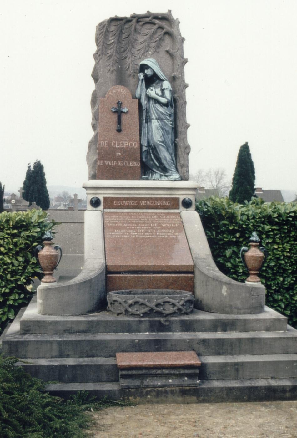 Grafmonument met een beeld van een treurige vrouw en een kruis, omgeven door groen en vazen.