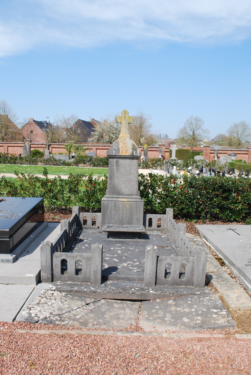 Oud grafmonument met kruis en omlijsting van stenen hekjes op een begraafplaats bij zonnig weer.