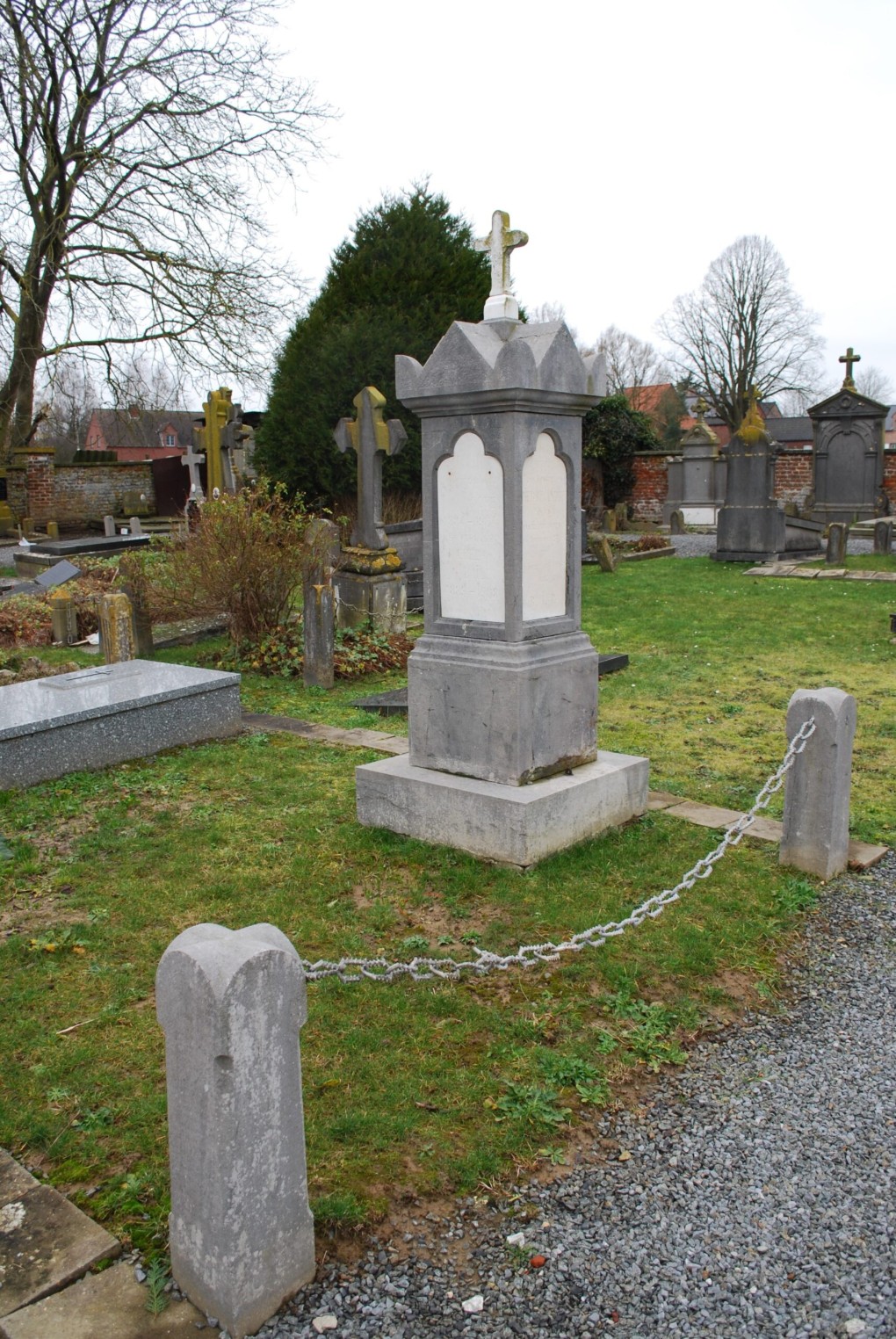 Begraafplaats met verschillende grafstenen en een centraal monument met een kruis erop, omgeven door gras en bomen.