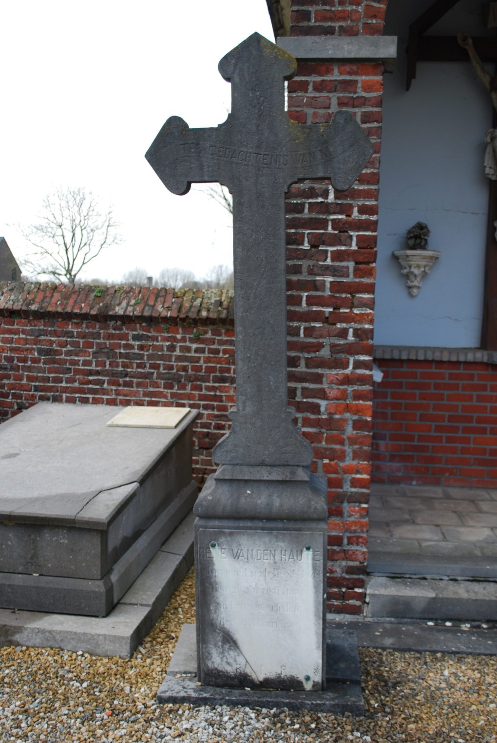 Grafsteen met kruis en inscriptie, geplaatst bij een bakstenen muur en een grafkelder.