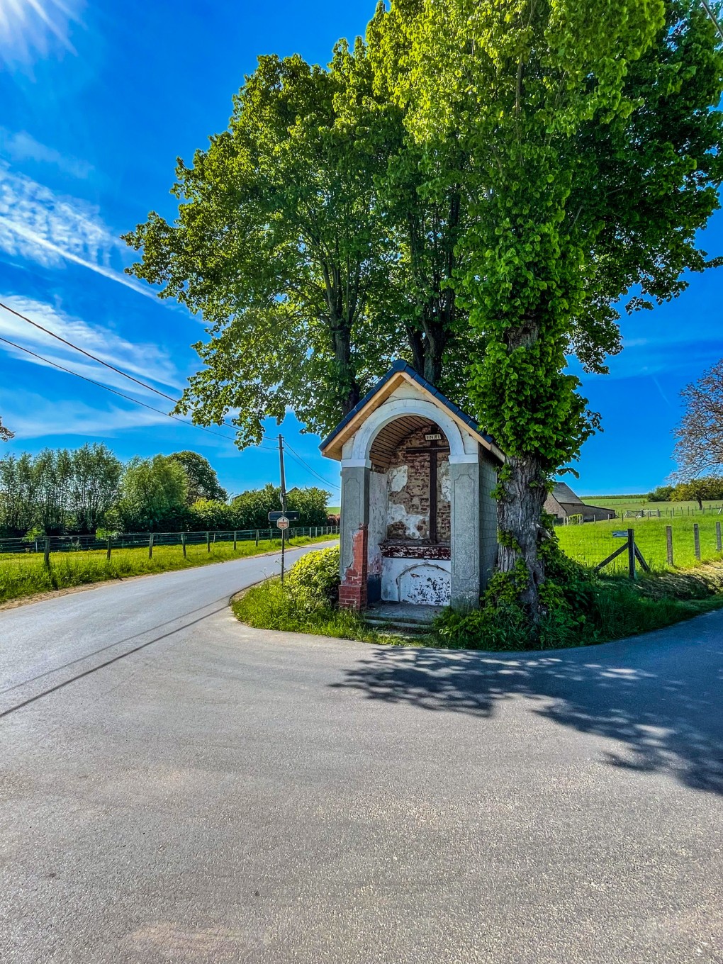 Kleine kapel met kruis bij een boom op een kruispunt in een groene, landelijke omgeving onder een blauwe hemel.