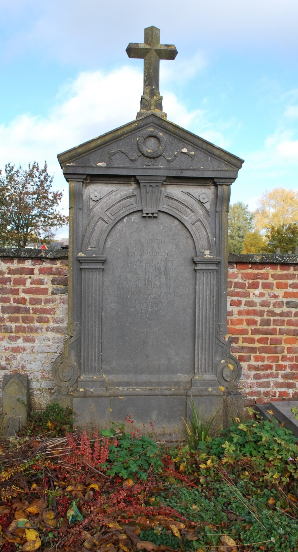 Oud grafsteen met kruis en versieringen, omringd door herfstbladeren en een bakstenen muur.