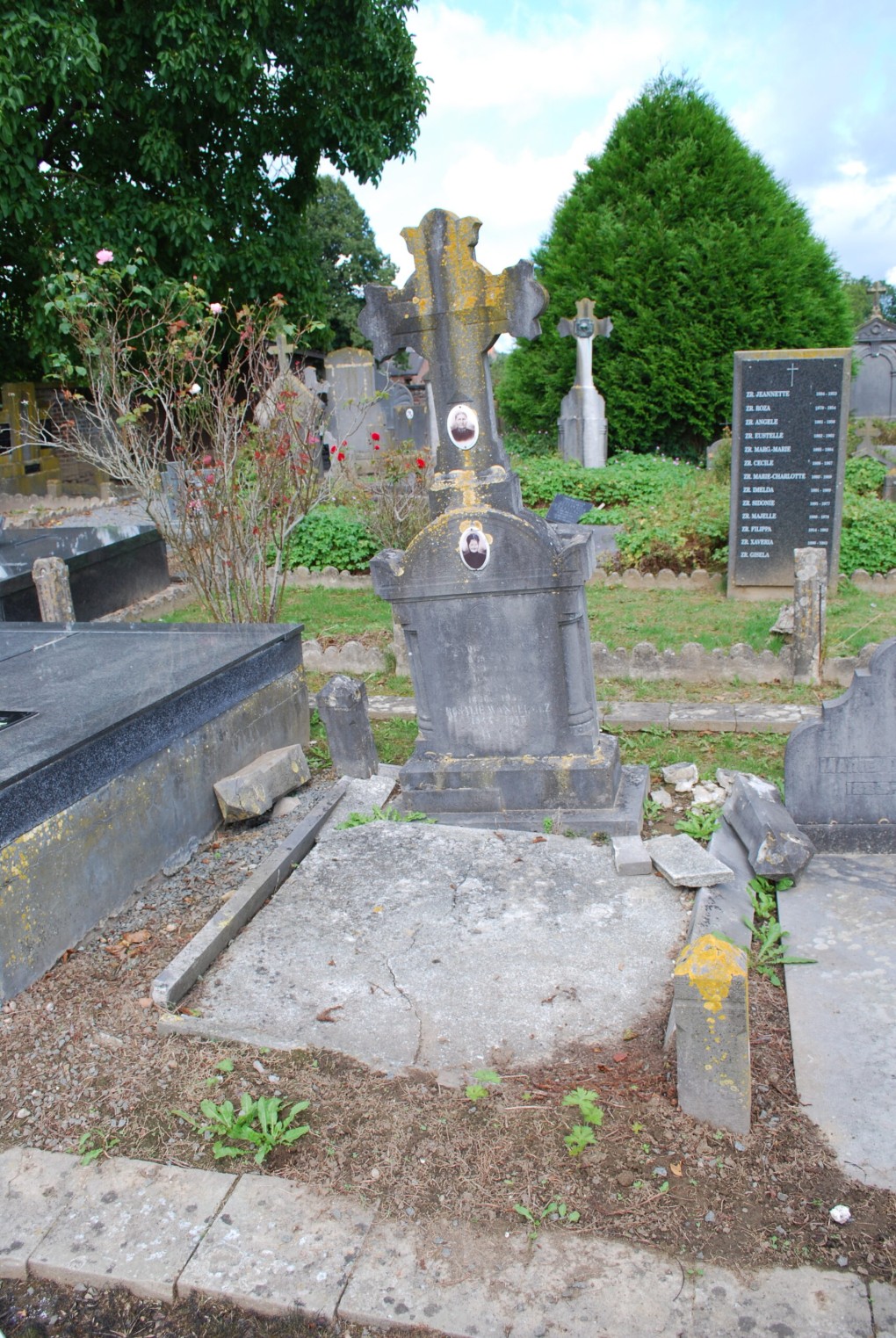 Oude grafsteen met kruis en foto's op een begraafplaats omringd door groen en andere graven.