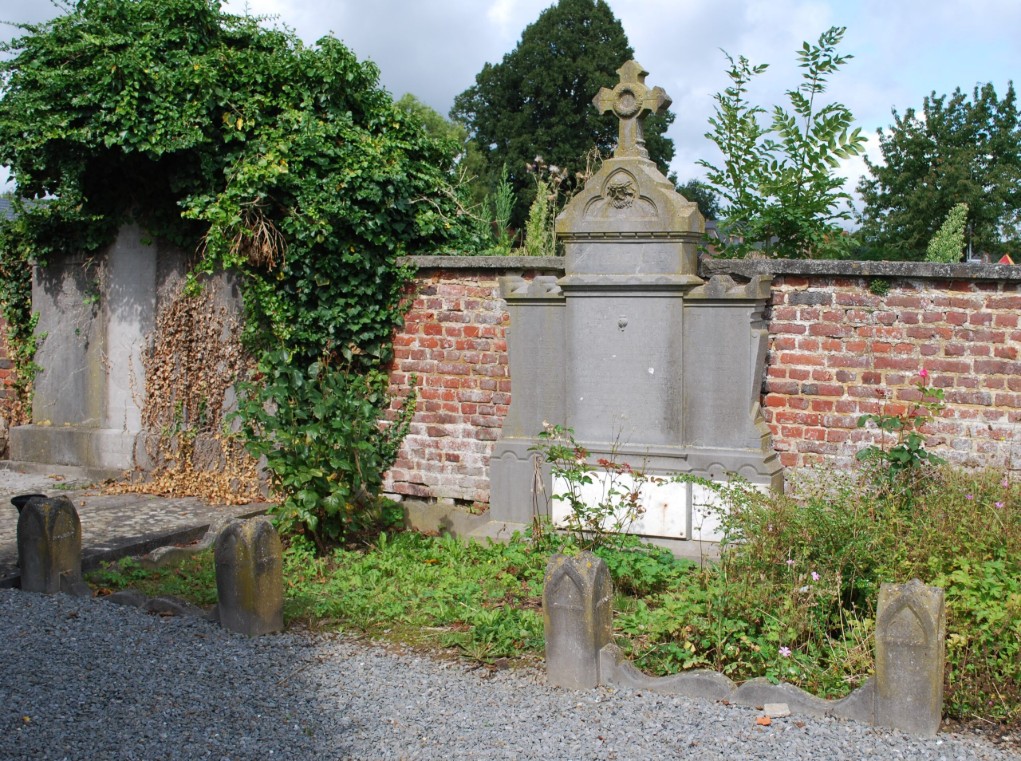 Oud grafmonument met kruis bij een bakstenen muur, omringd door groen en klimop.