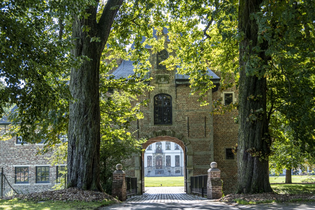 Oude poort van Lilare met bakstenen muur en grote bomen aan weerszijden, zonlicht schijnt door het groen.
